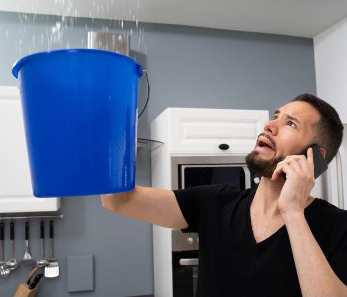 Man standing in kitchen with bucket catching water pouring from the second floor while on the phone with his insurance
